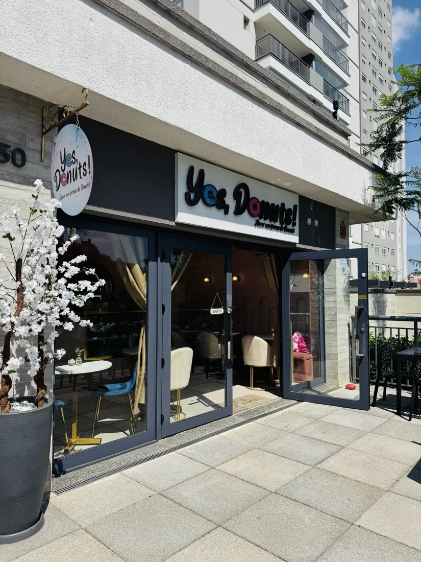 Fachada da Yes, Donuts! na Rua Celso Ramos, 150, com letreiro, portas de vidro e árvore de flores brancas na entrada